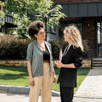 blonde real estate agent talking to african american client near house and for sale signboard Makler Grevenbroich, Hausverkauf, Immobilien, Maklerin spricht mit Kundin, Im Hintergrund ist ein Haus zu sehen